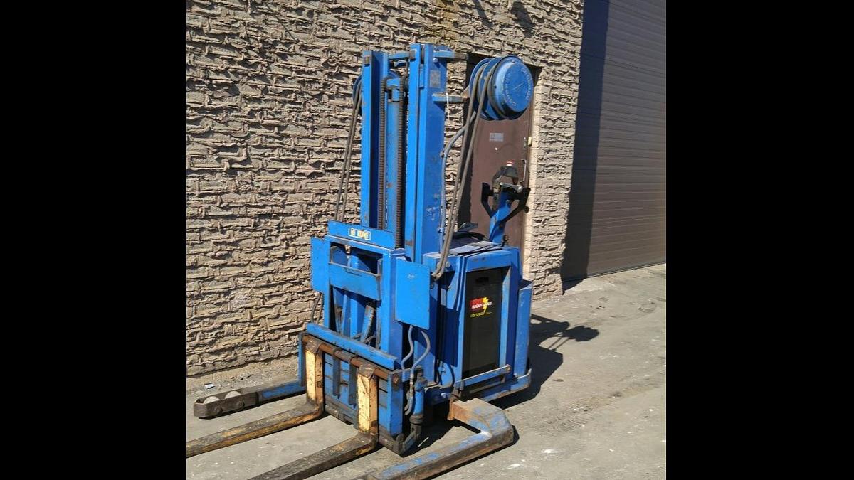 Front angled view of refurbished 1995 Blue Giant NA120W30 electric walkie straddle stacker reach showing straddle legs, mast, 42" forks, battery compartment, and CSA certification decal during Brampton deployment.