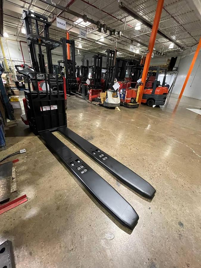 Angled opposite side view of refurbished 2014 Raymond 8410 electric rider pallet truck showing drive unit cover, platform entry, and fork tips during Toronto deployment